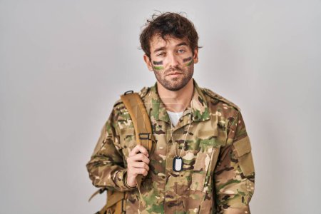 Foto de Joven hispano vistiendo uniforme camuflaje del ejército con aspecto de sueño y cansado, agotado por la fatiga y la resaca, ojos perezosos en la mañana. - Imagen libre de derechos