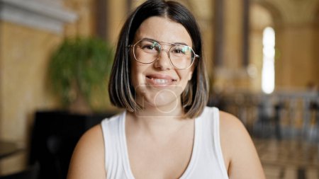 Foto de Joven mujer hispana hermosa con gafas sonriendo feliz sentado en la mesa en la cafetería - Imagen libre de derechos