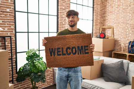 Photo for Arab man with beard holding welcome doormat smiling looking to the side and staring away thinking. - Royalty Free Image