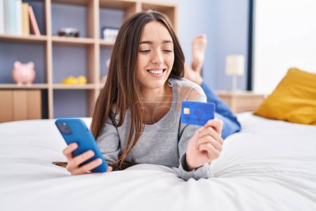 Photo for Young beautiful hispanic woman using smartphone and credit card lying on bed at bedroom - Royalty Free Image