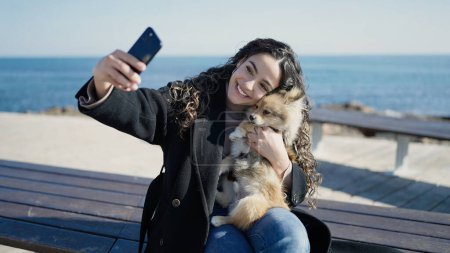 Foto de Mujer hispana joven con perro sonriendo confiado hacer selfie por teléfono inteligente en la playa - Imagen libre de derechos