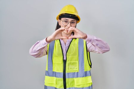 Foto de Chica hispana vistiendo uniforme de constructor y hardhat sonriendo en el amor haciendo forma de símbolo del corazón con las manos. concepto romántico. - Imagen libre de derechos