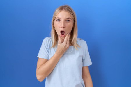 Foto de Mujer caucásica joven con camiseta azul casual que se ve fascinada con la incredulidad, sorpresa y expresión asombrada con las manos en la barbilla - Imagen libre de derechos