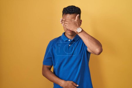 Photo for Young hispanic man standing over yellow background covering eyes with hand, looking serious and sad. sightless, hiding and rejection concept - Royalty Free Image