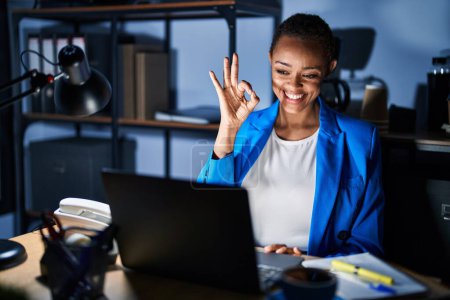 Foto de Hermosa mujer afroamericana que trabaja en la oficina por la noche sonriendo positiva haciendo signo bien con la mano y los dedos. expresión exitosa. - Imagen libre de derechos