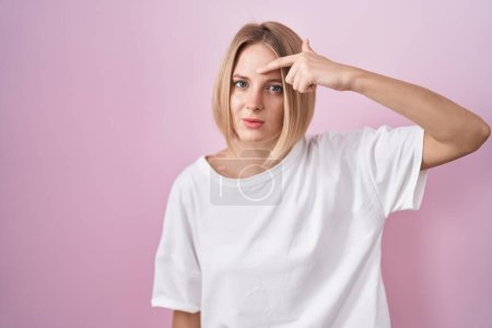 Téléchargez les photos : Young caucasian woman standing over pink background pointing unhappy to pimple on forehead, ugly infection of blackhead. acne and skin problem - en image libre de droit