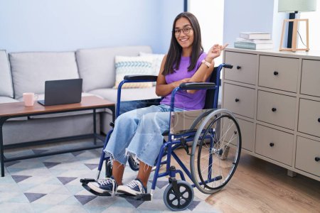 Photo for Young hispanic woman sitting on wheelchair at home with a big smile on face, pointing with hand and finger to the side looking at the camera. - Royalty Free Image