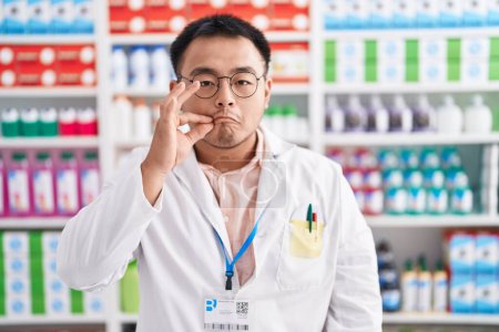 Téléchargez les photos : Jeune homme chinois travaillant à la pharmacie drugstore bouche et lèvres fermées comme zip avec les doigts. secret et silencieux, tabou parlant - en image libre de droit