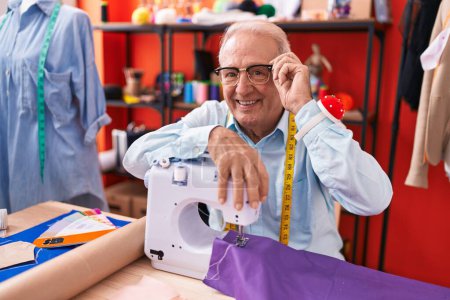 Foto de Hombre de pelo gris de mediana edad sastre sonriendo confiado apoyándose en la máquina de coser en el taller - Imagen libre de derechos