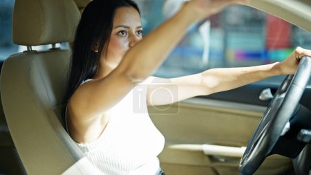 Photo for Young beautiful hispanic woman driver touching rearview sitting on car at street - Royalty Free Image