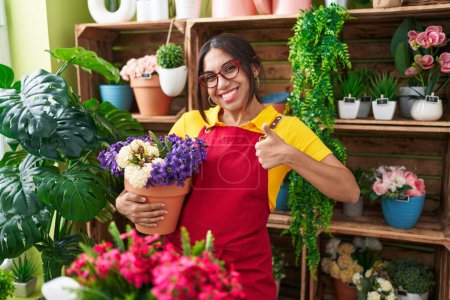 Téléchargez les photos : Jeune femme arabe travaillant au magasin de fleuriste tenant l'usine souriant heureux et positif, pouce vers le haut faisant l'excellent et signe d'approbation - en image libre de droit