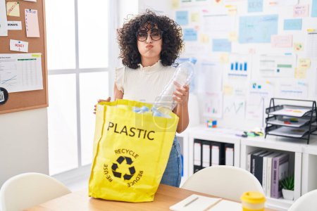 Téléchargez les photos : Jeune femme du Moyen-Orient tenant un sac de recyclage avec des bouteilles en plastique au bureau gonflant les joues avec un visage drôle. bouche gonflée d'air, attrapant l'air. - en image libre de droit