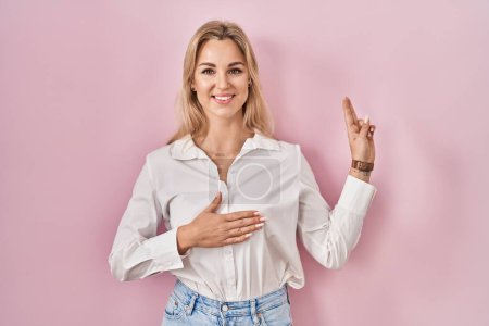 Foto de Mujer caucásica joven con camisa blanca casual sobre fondo rosa sonriendo jurando con la mano en el pecho y los dedos hacia arriba, haciendo una promesa de lealtad juramento - Imagen libre de derechos