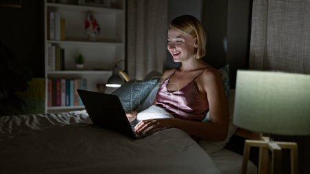 Photo for Young blonde woman using laptop sitting on bed smiling at bedroom - Royalty Free Image