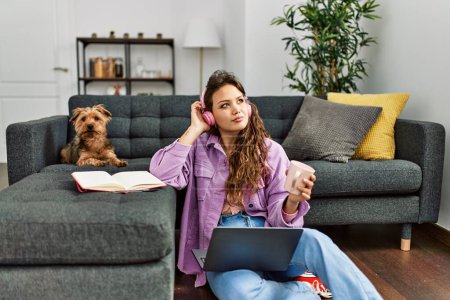 Photo for Young beautiful hispanic woman listening to music sitting on floor with dog at home - Royalty Free Image