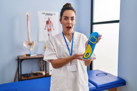 Photo for Young hispanic woman holding shoe insole at physiotherapy clinic afraid and shocked with surprise and amazed expression, fear and excited face. - Royalty Free Image