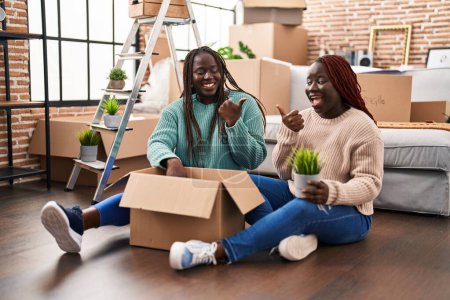 Téléchargez les photos : Deux femmes africaines se déplaçant vers une nouvelle maison assise sur le sol pointant le pouce vers le côté souriant heureux avec la bouche ouverte - en image libre de droit