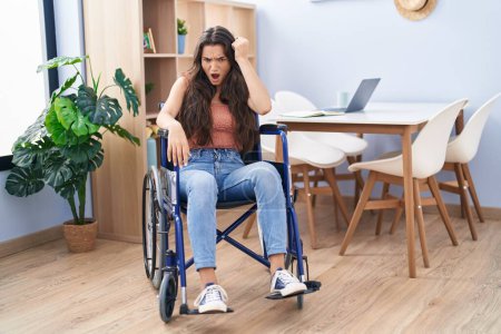 Photo for Young teenager girl sitting on wheelchair at the living room annoyed and frustrated shouting with anger, yelling crazy with anger and hand raised - Royalty Free Image