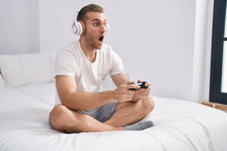 Téléchargez les photos : Jeune homme caucasien assis sur le lit à la maison jouer à des jeux vidéo peur et choqué avec surprise et expression étonnante, peur et visage excité. - en image libre de droit