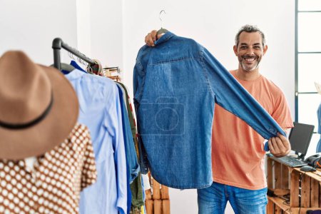Foto de Hombre de pelo gris de mediana edad cliente sonriendo confiado sosteniendo la ropa de rack en la tienda de ropa - Imagen libre de derechos