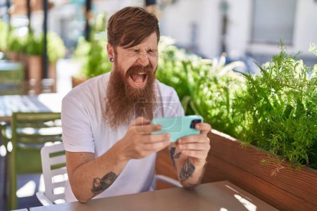 Téléchargez les photos : Young redhead man watching video on smartphone sitting on table at coffee shop terrace - en image libre de droit