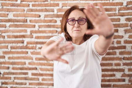 Foto de Mujer mayor con gafas de pie sobre la pared de ladrillos haciendo marco con las manos palmas y los dedos, perspectiva de la cámara - Imagen libre de derechos