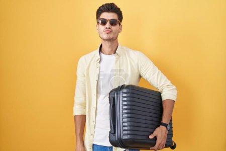 Foto de Joven hombre hispano sosteniendo maleta va de vacaciones de verano mirando a la cámara soplando un beso en el aire siendo encantador y sexy. expresión de amor. - Imagen libre de derechos