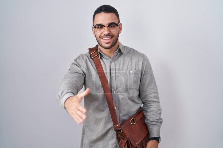 Foto de Joven hombre hispano con maleta sonriente amistoso ofreciendo apretón de manos como saludo y bienvenida. negocios exitosos. - Imagen libre de derechos