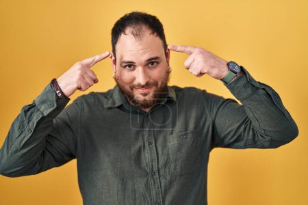 Photo for Plus size hispanic man with beard standing over yellow background smiling pointing to head with both hands finger, great idea or thought, good memory - Royalty Free Image