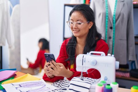 Téléchargez les photos : Jeune femme chinoise tailleur souriant confiant en utilisant smartphone à l'atelier - en image libre de droit