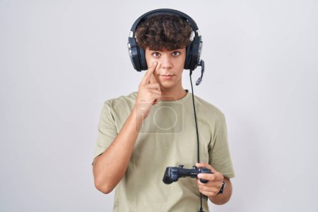 Photo for Hispanic teenager playing video game holding controller pointing to the eye watching you gesture, suspicious expression - Royalty Free Image