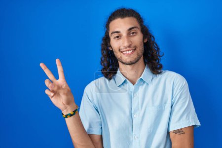 Téléchargez les photos : Jeune homme hispanique debout sur fond bleu souriant avec un sourire heureux clin d'oeil à la caméra faisant signe de victoire avec les doigts. numéro deux. - en image libre de droit