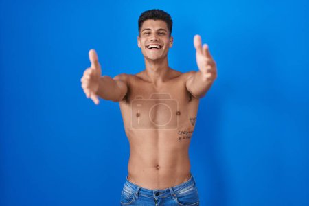 Photo for Young hispanic man standing shirtless over blue background looking at the camera smiling with open arms for hug. cheerful expression embracing happiness. - Royalty Free Image