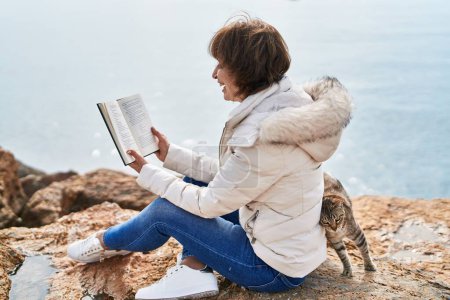 Photo for Middle age woman reading book sitting on the rock at seaside - Royalty Free Image