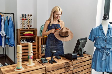 Foto de Mujer rubia joven asistente de tienda sonriente seguro de escaneo sombrero en la tienda de ropa - Imagen libre de derechos