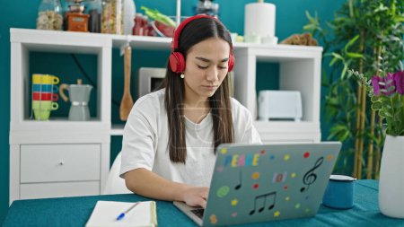 Téléchargez les photos : Jeune belle femme hispanique utilisant ordinateur portable et écouteurs assis sur la table à manger - en image libre de droit