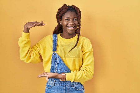 Foto de Mujer africana de pie sobre fondo amarillo gesticulando con las manos mostrando un signo de tamaño grande y grande, símbolo de medida. sonriendo mirando a la cámara. concepto de medición. - Imagen libre de derechos