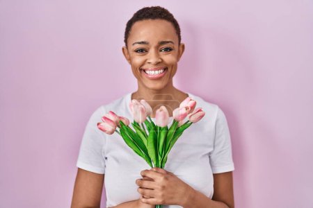 Foto de Hermosa mujer afroamericana sosteniendo ramo de flores de tulipanes rosados sonriendo con una sonrisa feliz y fresca en la cara. mostrando dientes. - Imagen libre de derechos