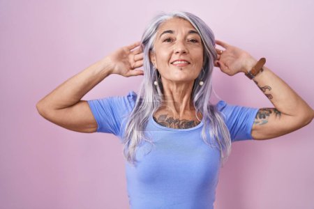 Foto de Mujer de mediana edad con tatuajes de pie sobre fondo rosa sonriendo tirando de las orejas con los dedos, gesto divertido. problema de audición - Imagen libre de derechos