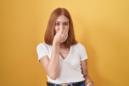 Téléchargez les photos : Jeune femme rousse debout sur fond jaune sentant quelque chose de puant et dégoûtant, odeur intolérable, retenant la respiration avec les doigts sur le nez. mauvaise odeur - en image libre de droit