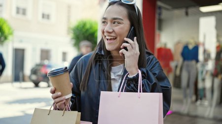 Foto de Joven hermosa mujer hispana hablando en el teléfono inteligente sosteniendo bolsas de compras y café en la calle - Imagen libre de derechos