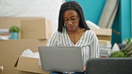 Téléchargez les photos : Femme afro-américaine utilisant un ordinateur portable assis sur le canapé à la nouvelle maison - en image libre de droit