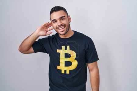 Téléchargez les photos : Jeune homme hispanique portant Bitcoin t-shirt souriant avec la main sur l'oreille à l'écoute d'une audience à la rumeur ou des commérages. concept de surdité. - en image libre de droit