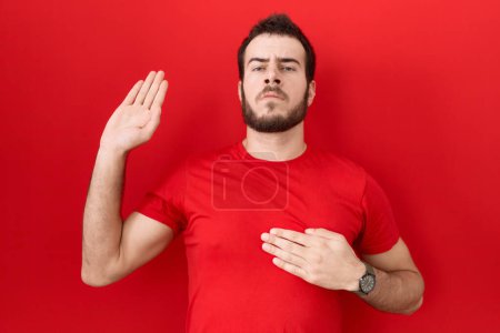 Téléchargez les photos : Jeune homme hispanique portant un t-shirt rouge décontracté jurant avec la main sur la poitrine et la paume ouverte, faisant un serment de fidélité - en image libre de droit