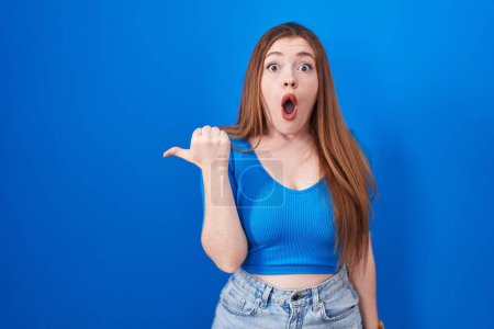 Photo for Redhead woman standing over blue background surprised pointing with hand finger to the side, open mouth amazed expression. - Royalty Free Image