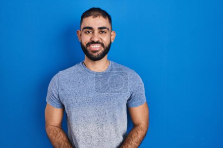 Photo for Middle east man with beard standing over blue background with hands together and crossed fingers smiling relaxed and cheerful. success and optimistic - Royalty Free Image
