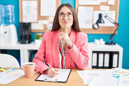 Photo for Young hispanic woman working at the office wearing glasses thinking concentrated about doubt with finger on chin and looking up wondering - Royalty Free Image