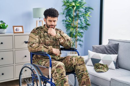 Photo for Arab man wearing camouflage army uniform sitting on wheelchair looking confident at the camera with smile with crossed arms and hand raised on chin. thinking positive. - Royalty Free Image
