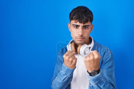 Téléchargez les photos : Young hispanic man standing over blue background ready to fight with fist defense gesture, angry and upset face, afraid of problem - en image libre de droit