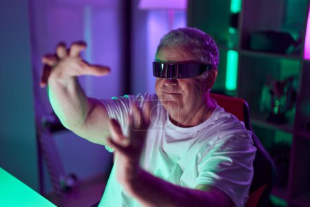 Téléchargez les photos : Homme aux cheveux gris moyen âge streamer jouer à un jeu vidéo en utilisant des lunettes de réalité virtuelle à la salle de jeux - en image libre de droit